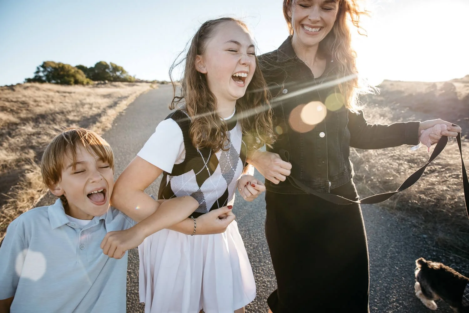 Single mom and two kids walk down path at Ring Mountain, laughing hard, with fields of golden grass and sunset light behind them. Their pet dog walks next to them. From single parent family photo shoot.