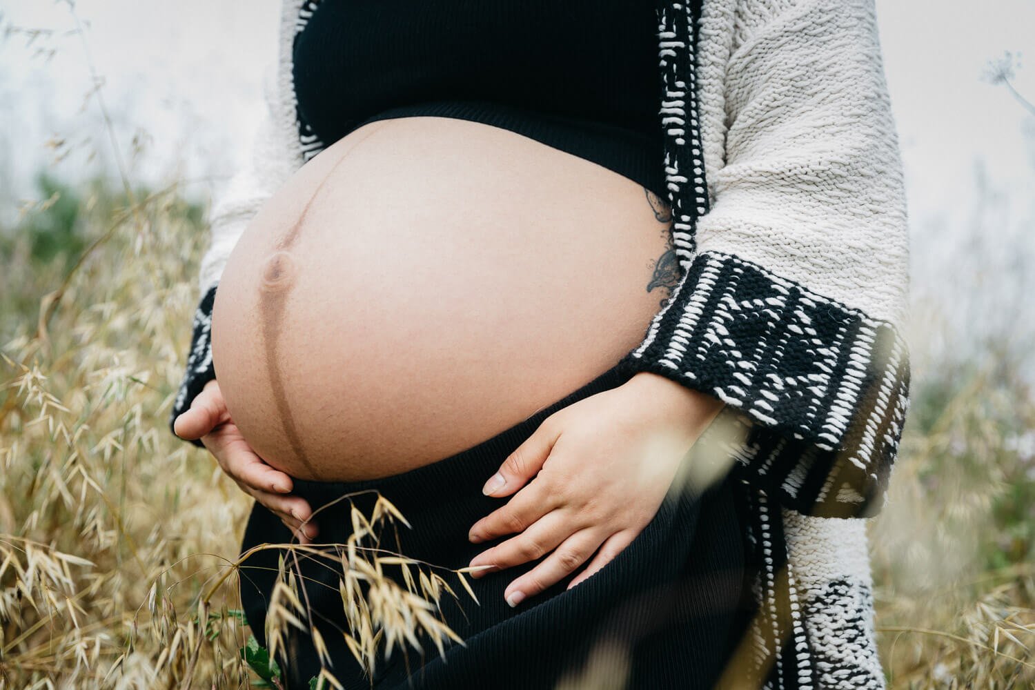 Pregnant woman shows exposed belly bump in field of long grasses during outdoor maternity session in Berkeley Marina.