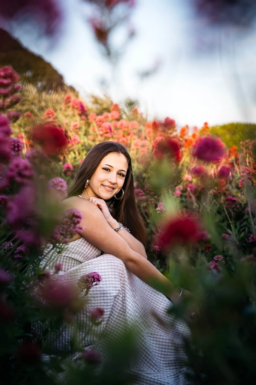 San Francisco senior photographer photographs high school senior in a white gingham dress sitting among vibrant pink wildflowers at the Albany Bulb in warm golden hour light.