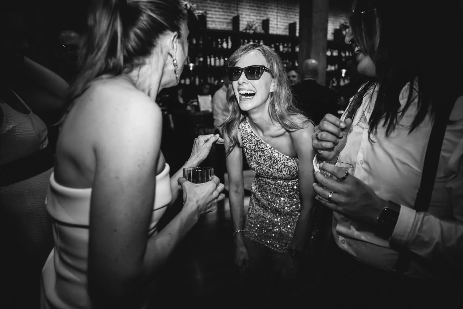 Woman in sunglasses and sequinned dress laughs with friends at bar, while flash lights up their faces.