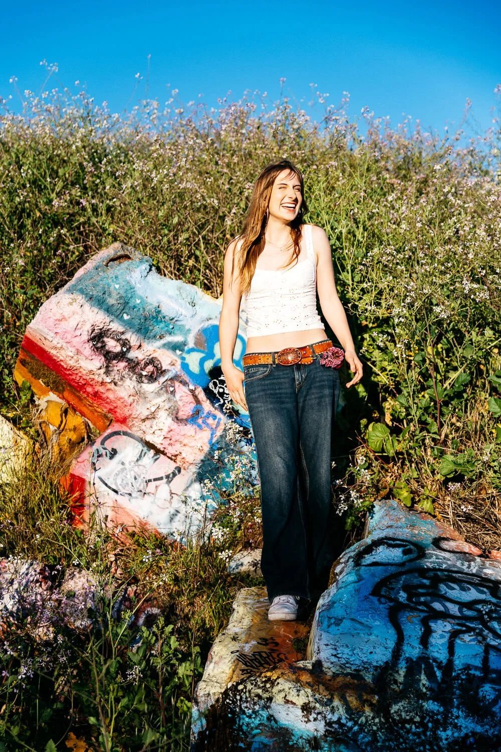 Berkeley senior photographer photographs high school senior laughing freely beside colorful graffiti-covered rocks at the Albany Bulb, wearing a white tank top, wide-leg jeans, and a western belt.