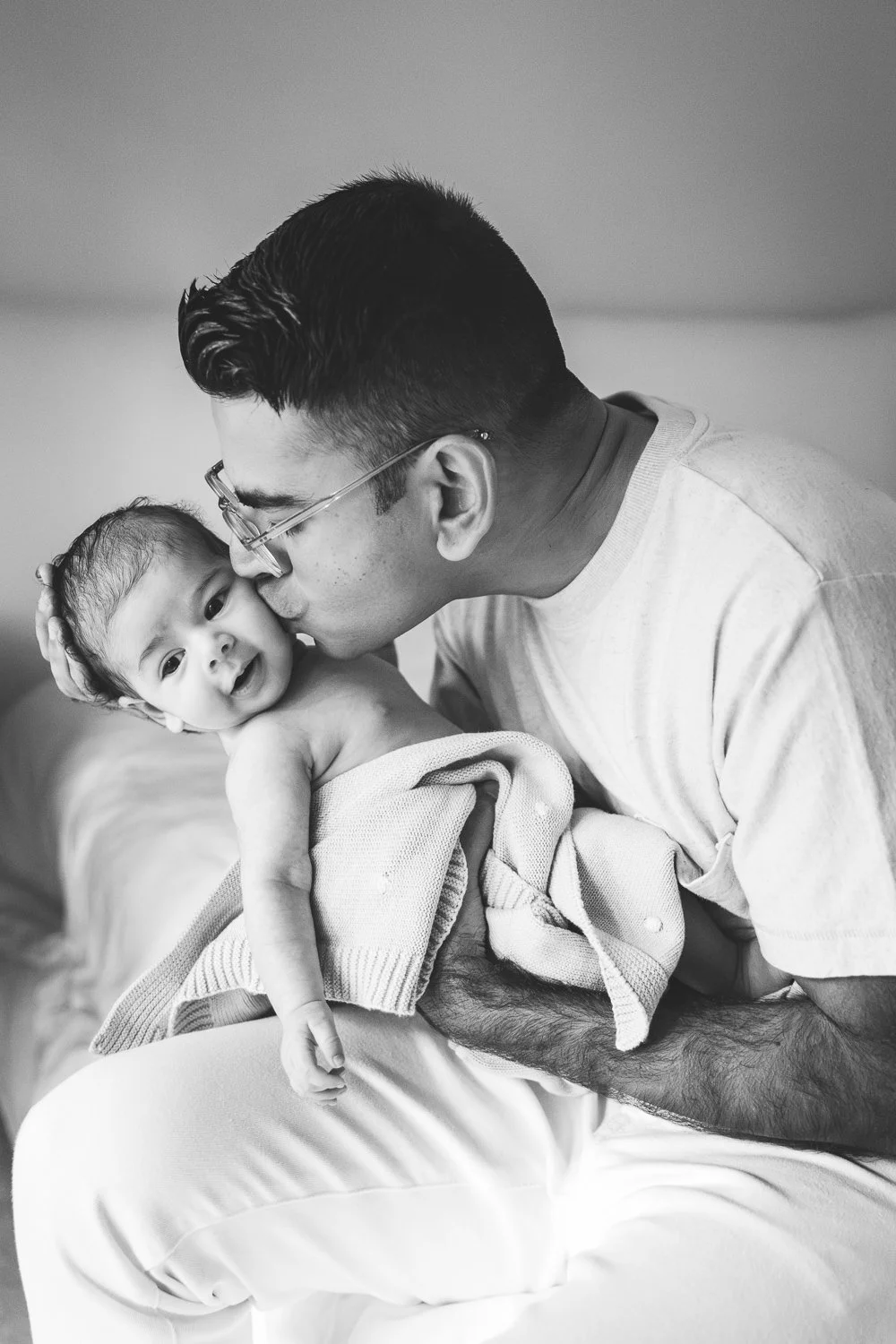 Dad snuggles and kisses newborn baby, who is gazing at camera.