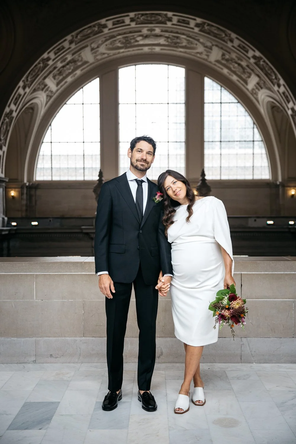 San Francisco City Hall elopement portrait with pregnant bride in white dress. The couple is posed to highlight their connection, with the bride leaning into her husband with head on shoulder. They hold hands.
