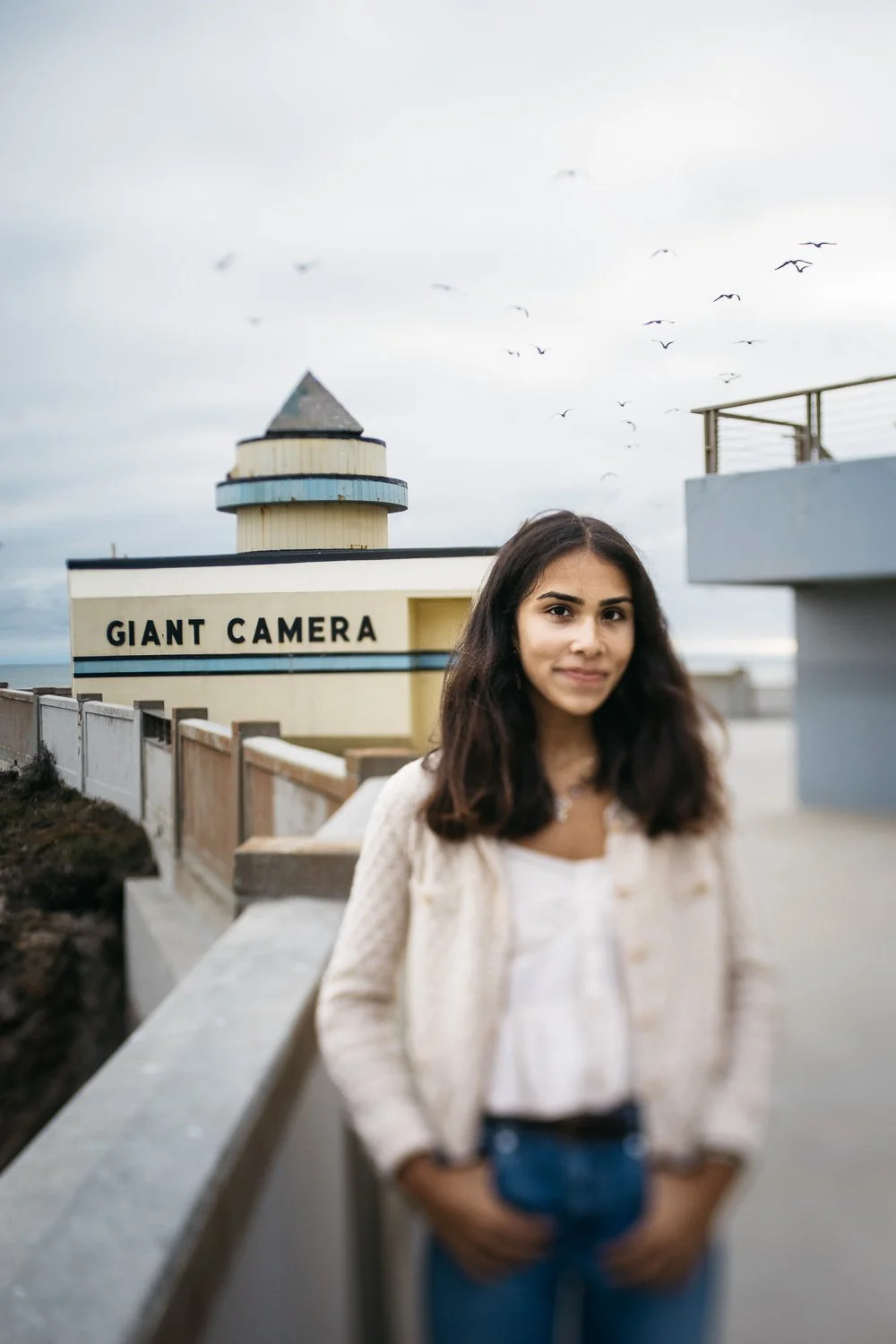 Teen girl wearing crea sweater and jeans stands with hands in pockets in front of the Giant Camera at the Cliffhouse.