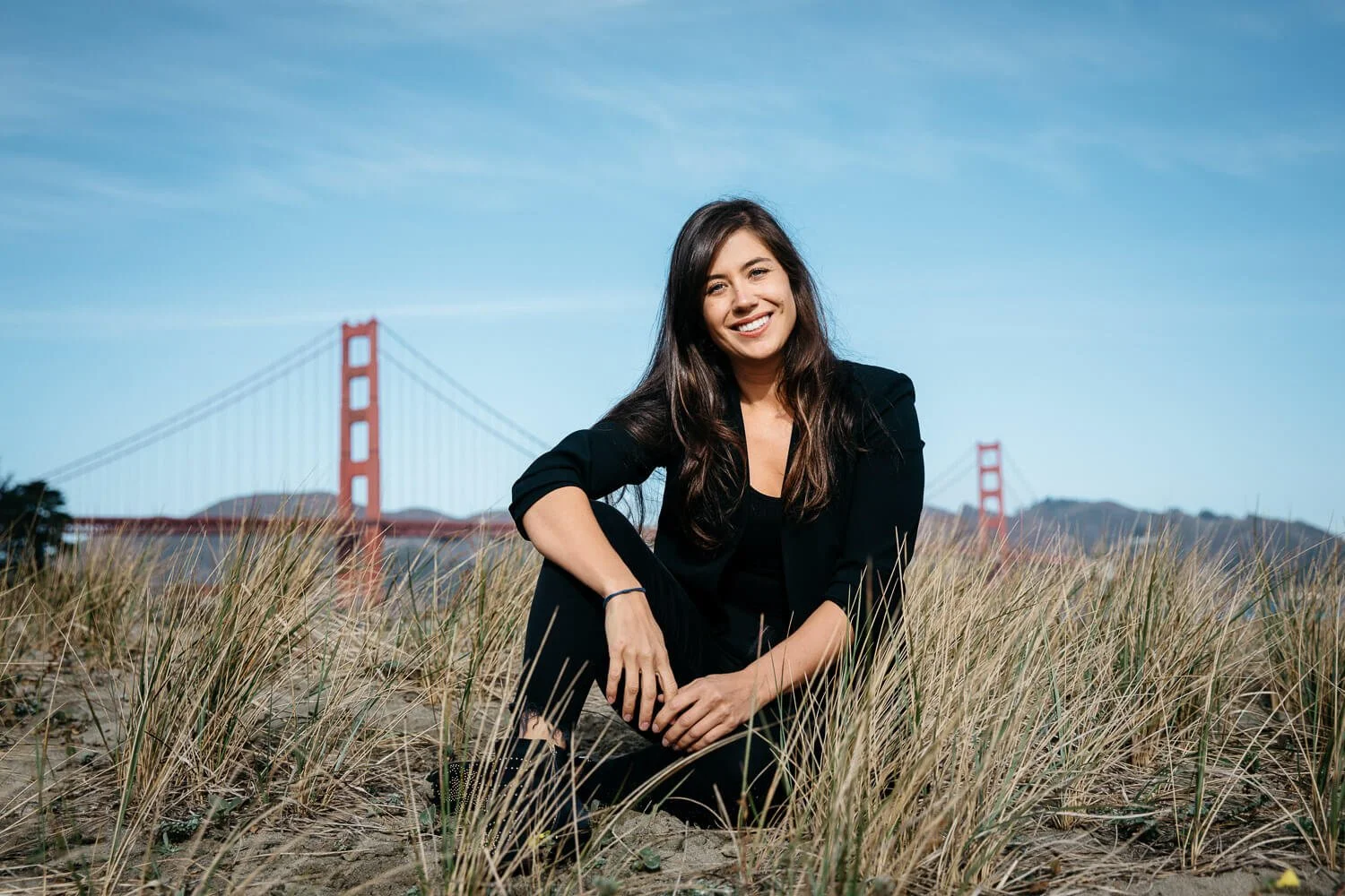 Dating by Blaine matchmaker, Tara Bratt, sits in dune grass at Crissy Field with the Golden Gate Bridge in the background.