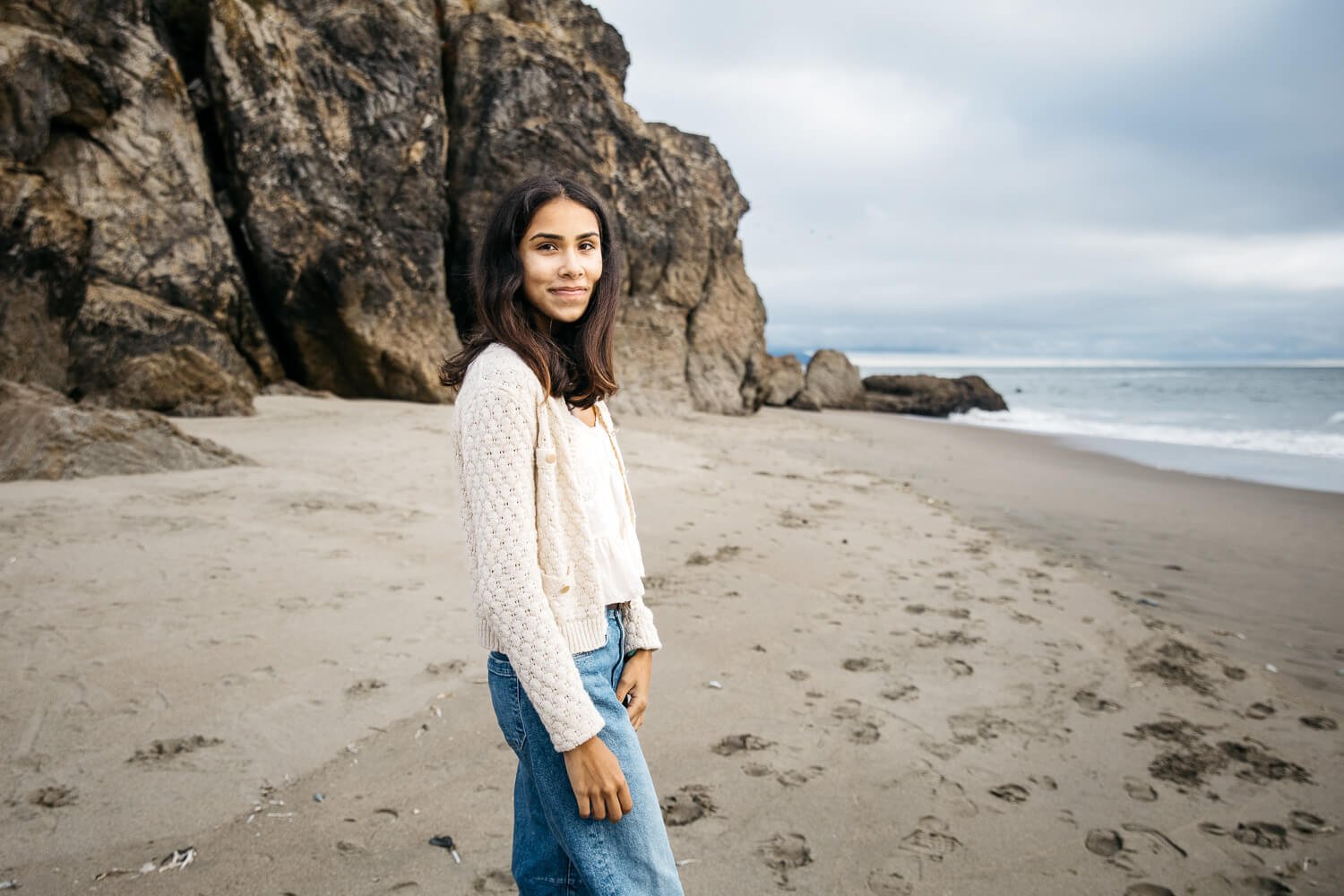 High school senior girl in white sweater stands in front of cliffs at Ocean Beach.