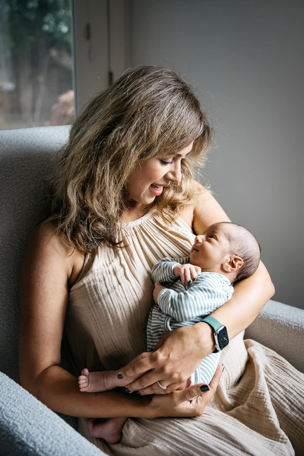 Mom sits in white chair next to window, holding newborn baby in her arms. She looks at her infant lovingly and he looks back at her with wide eyes. Taken during in home newborn lifestyle photo shoot in San Francisco.