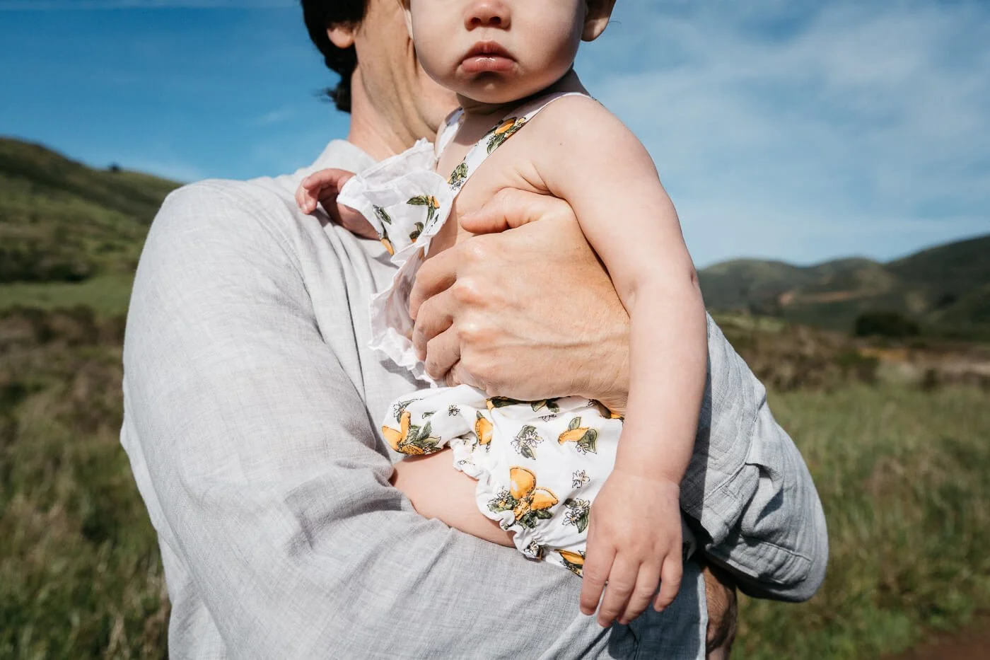 Crop portrait of the bottom of a baby's face and her torso, being held in dad's arms. Shot in the Marin Headlands during a family session.