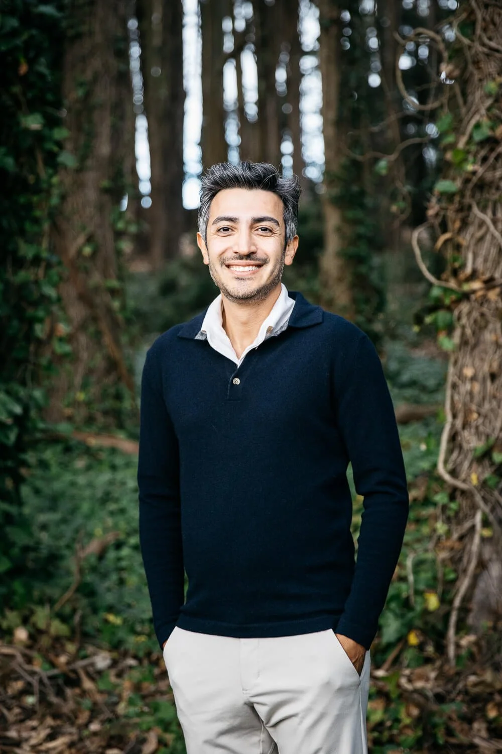 Man with dark hair and navy sweater stands smiling in eucalyptus grove in the Presidio in San Francisco. 