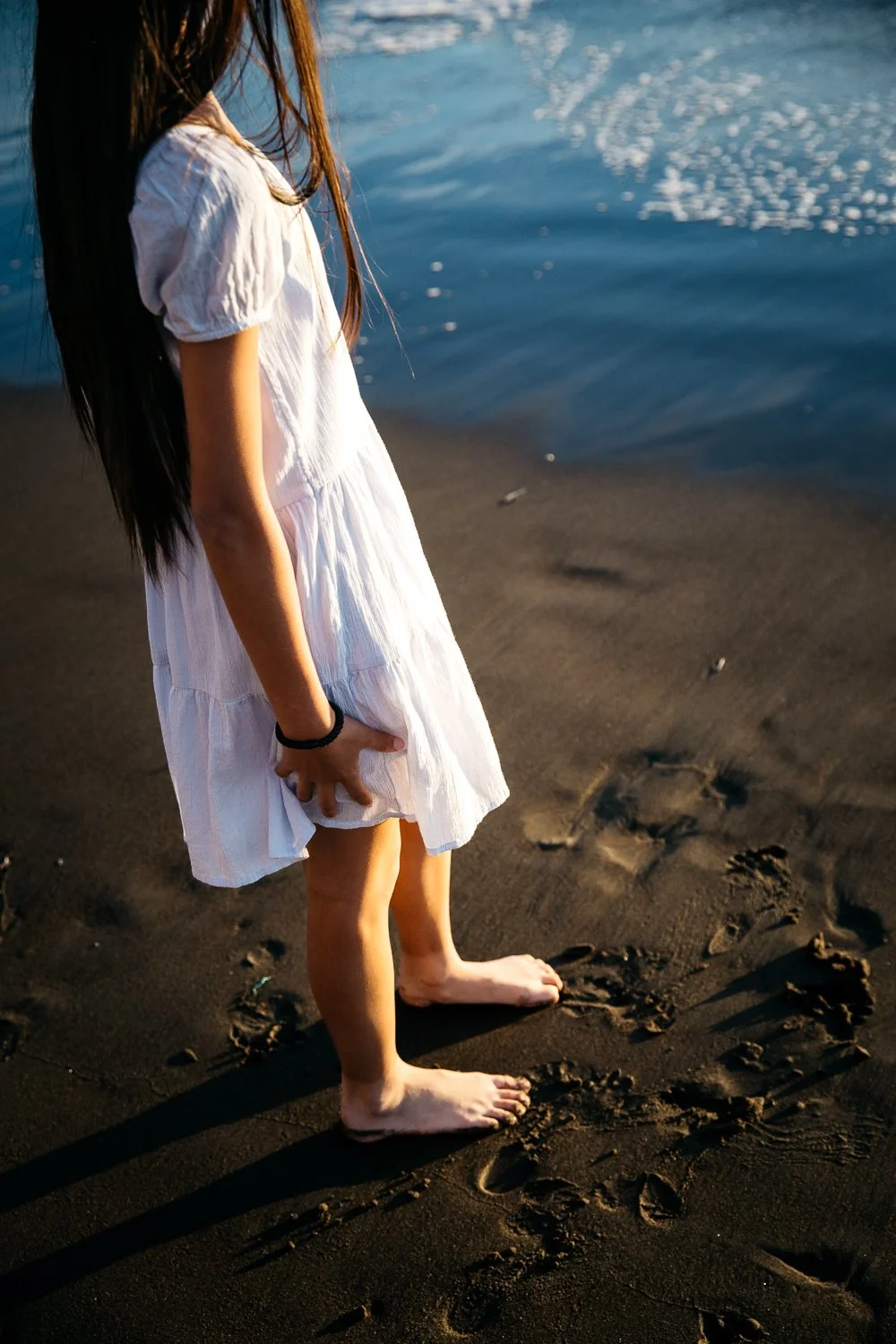 Over the shoulder shot of girl in white cotton dress stands in the surf at Rodeo Beach.