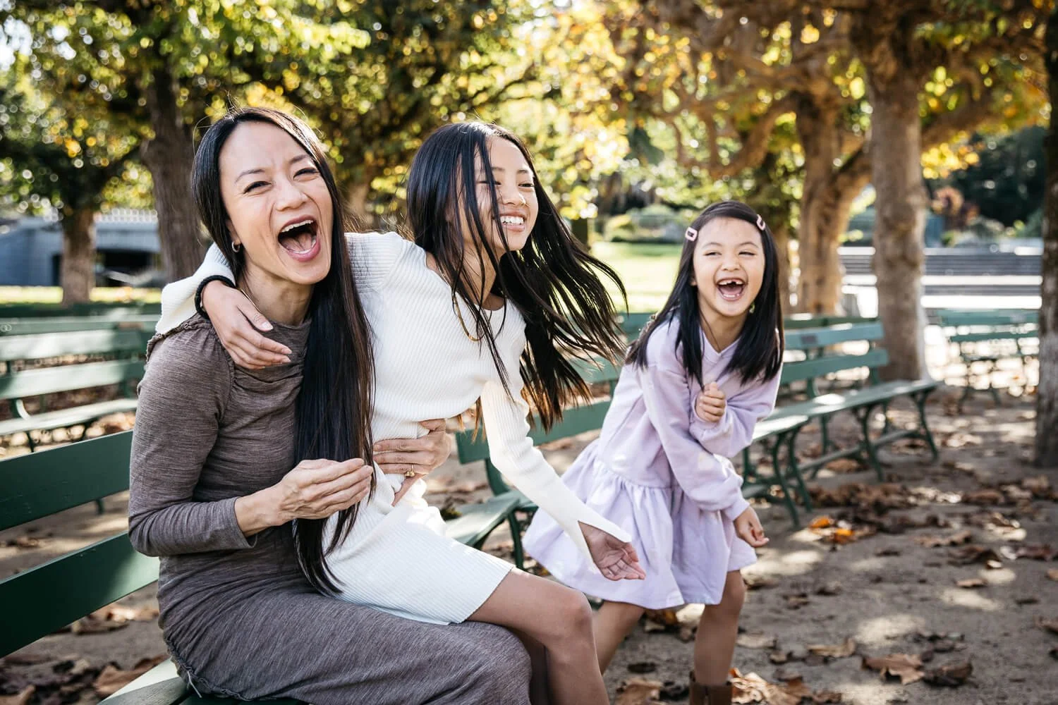 Single mom and 2 daughters sit on green benches in Golden Gate Park, laughing wildly together.