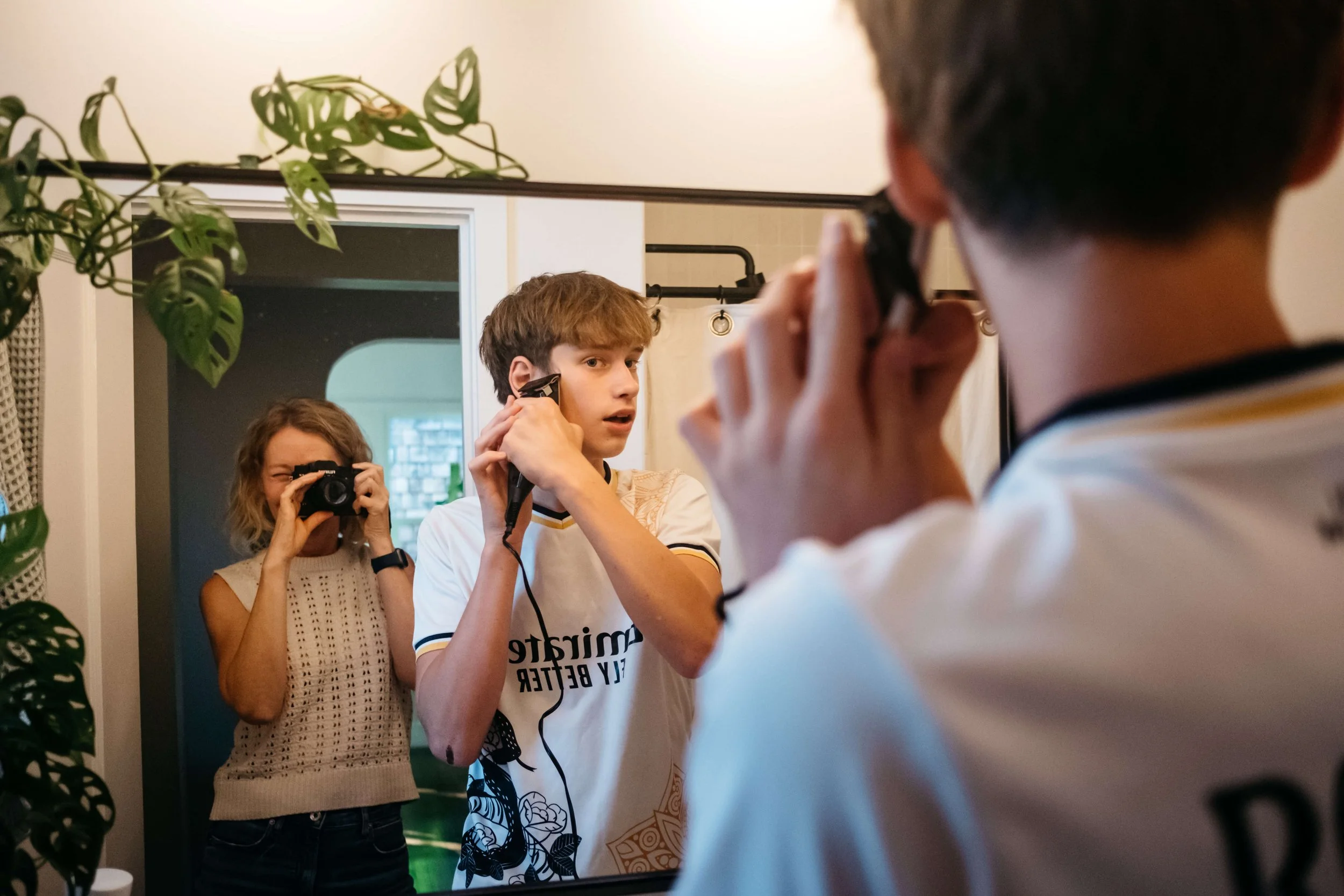 Teen boy looks in mirror while holding hair clippers up to head, while mom takes photo in the background.