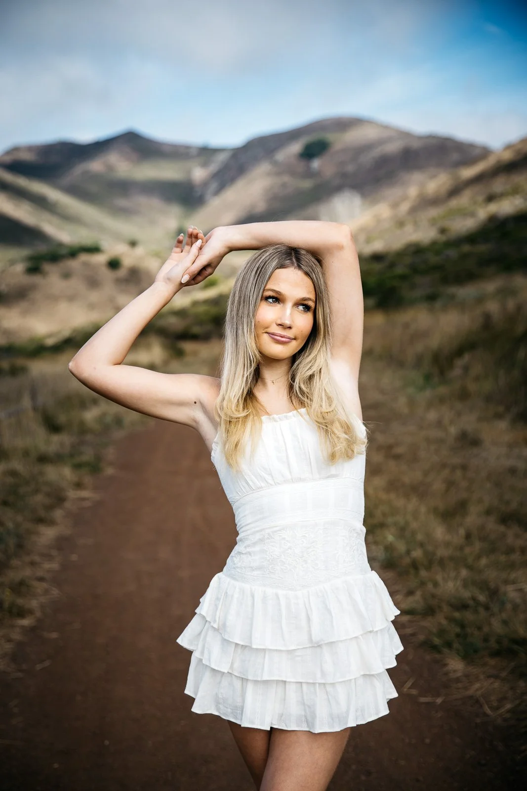 Teen girl with blond hair and white, ruffled dress stands with her arms overhead, shot during senior photo shoot in the Marin Headlands.