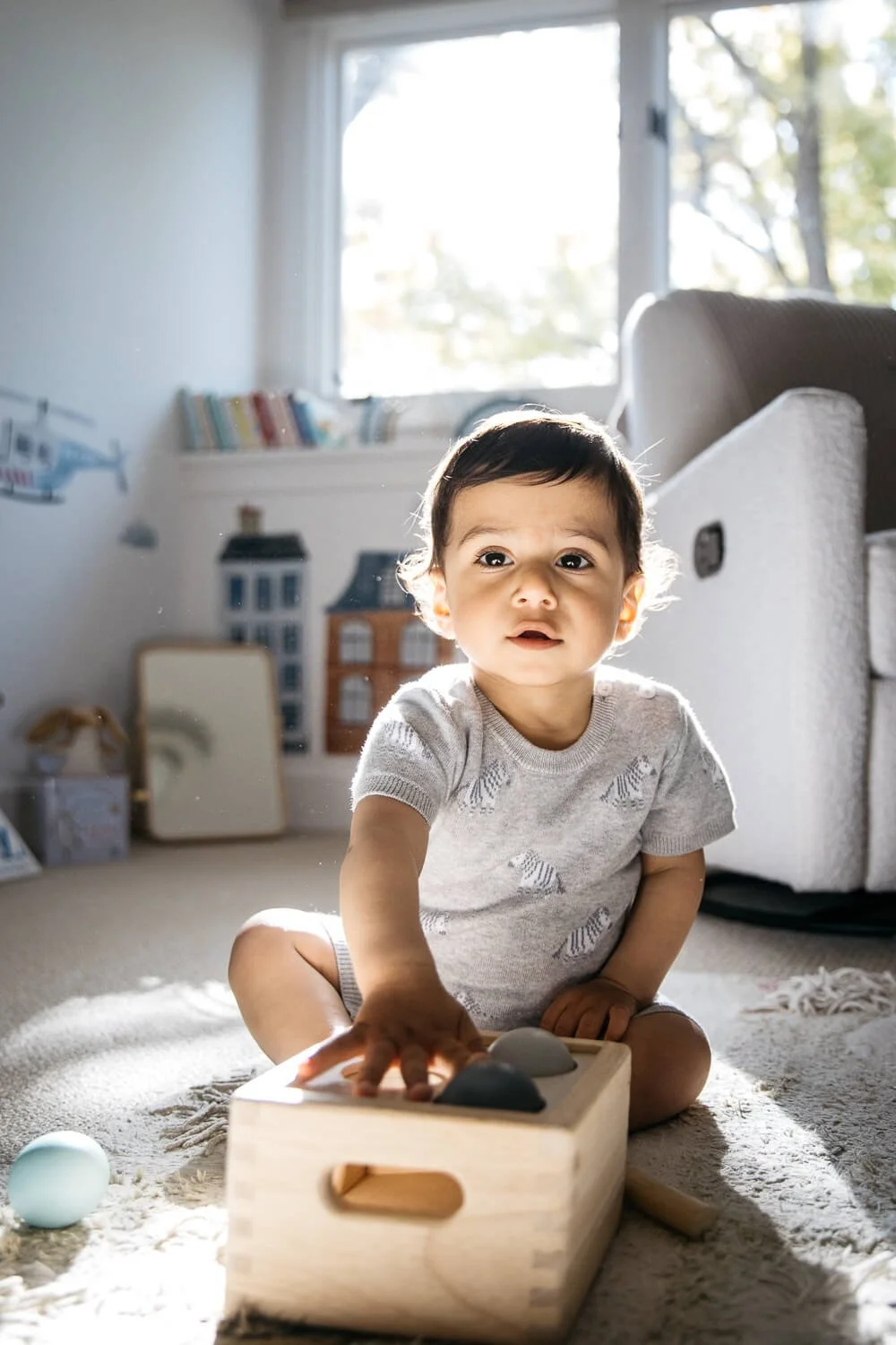 Baby in gray romper plays with wooden toys in light colored room, with bright sunlight from window backlighting him.