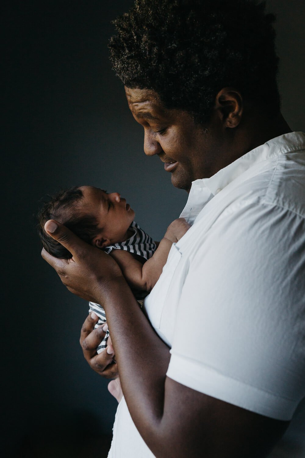Profile of Black dad in white shirt, gazing with wonder at newborn baby daughter in his arms.