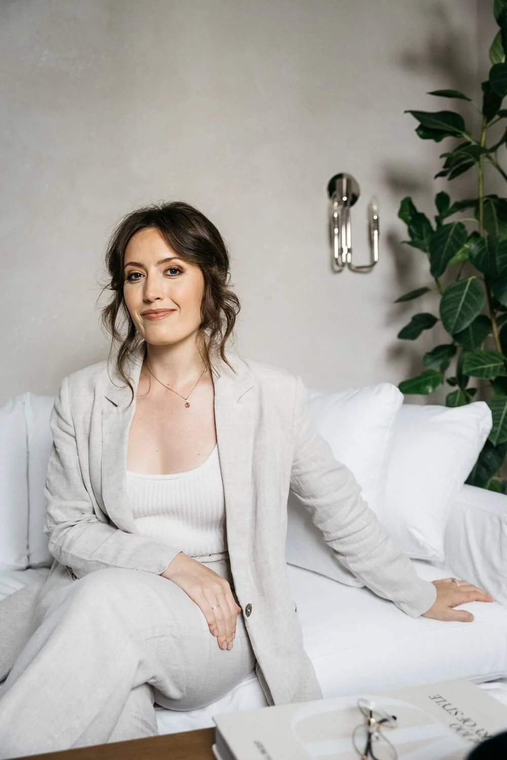 Brunette woman posed sitting confidently on white couch during a brand portrait session in Berkeley. Her posture is soft and natural, with one arm draped over her legs. She looks directly into camera with a small smile to give off easy confidence.