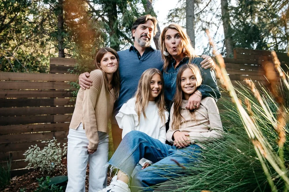 Mom, Dad, and three daughter stand in group hug and make silly faces at the camera, while standing in their backyard with trees, green grasses and a wooden fence.