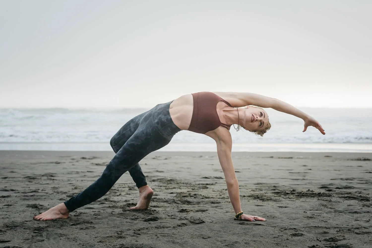 Yogi in gray leggings and marron top poses in front of water at Ocean Beach.