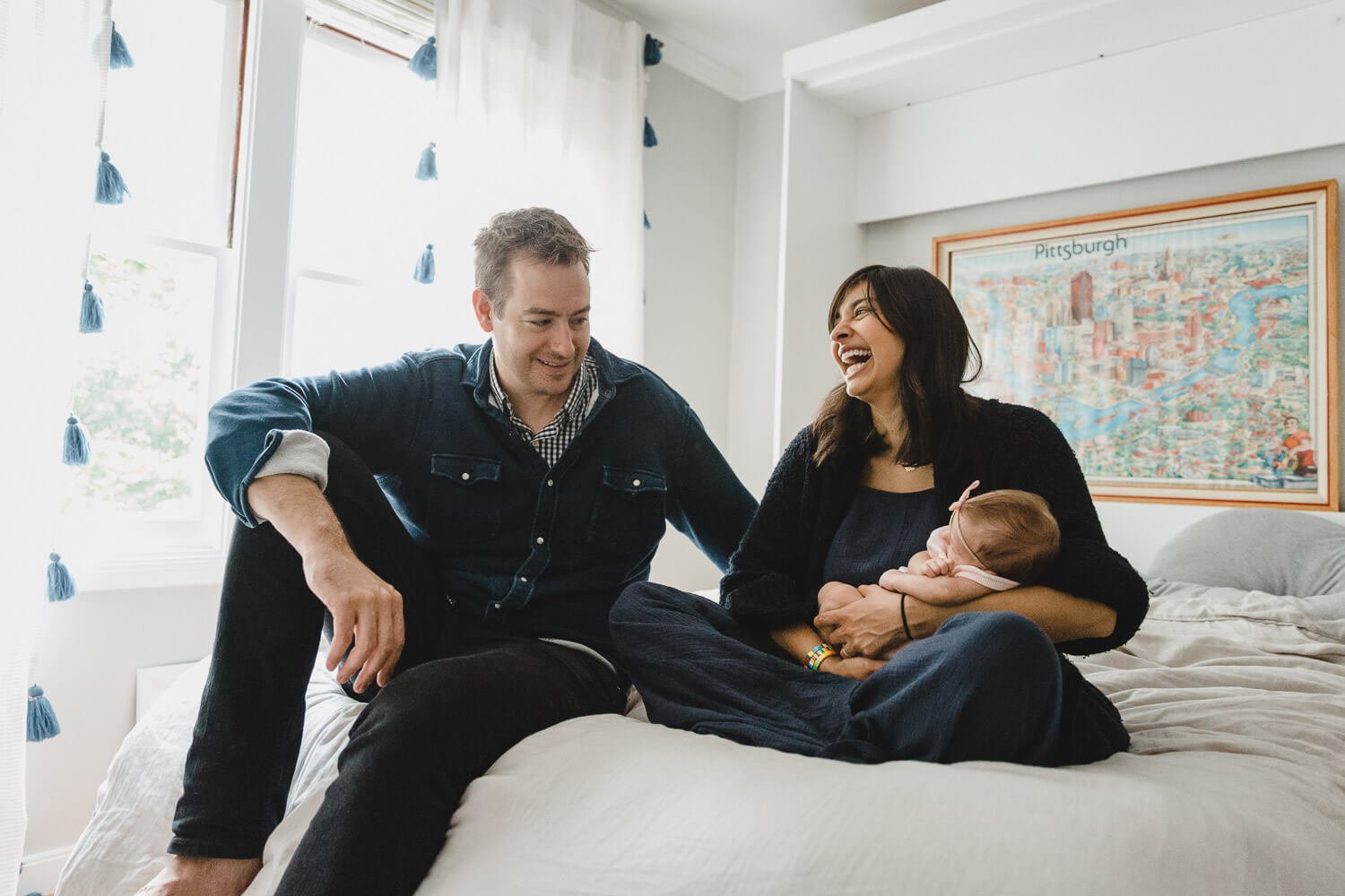Mom and dad look at each and laugh while sitting on bed with white cover and holding newborn in arms.