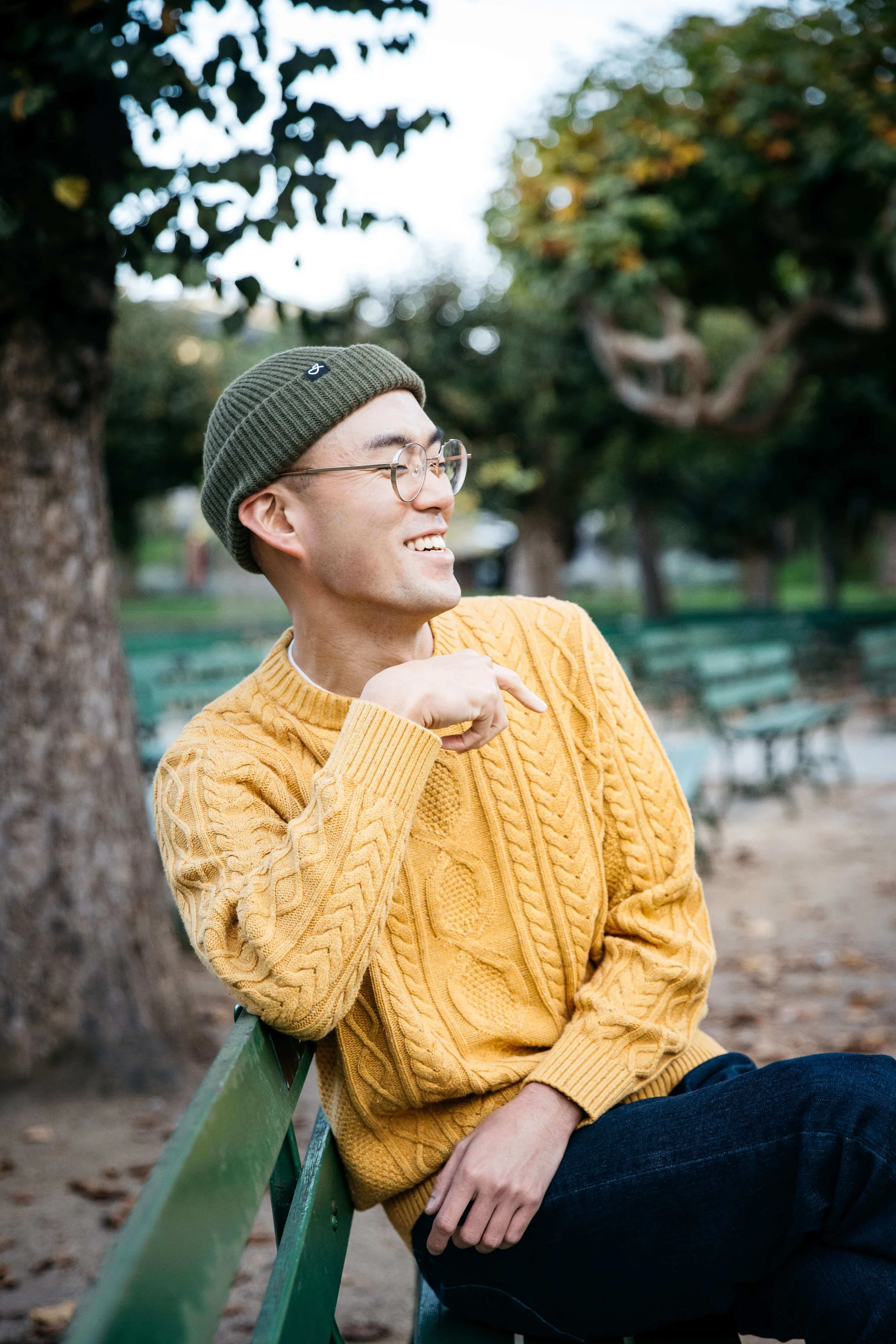 Asian man in yellow sweater and green beanie hat sits on green bench and laughs, shot in Golden Gate Park.