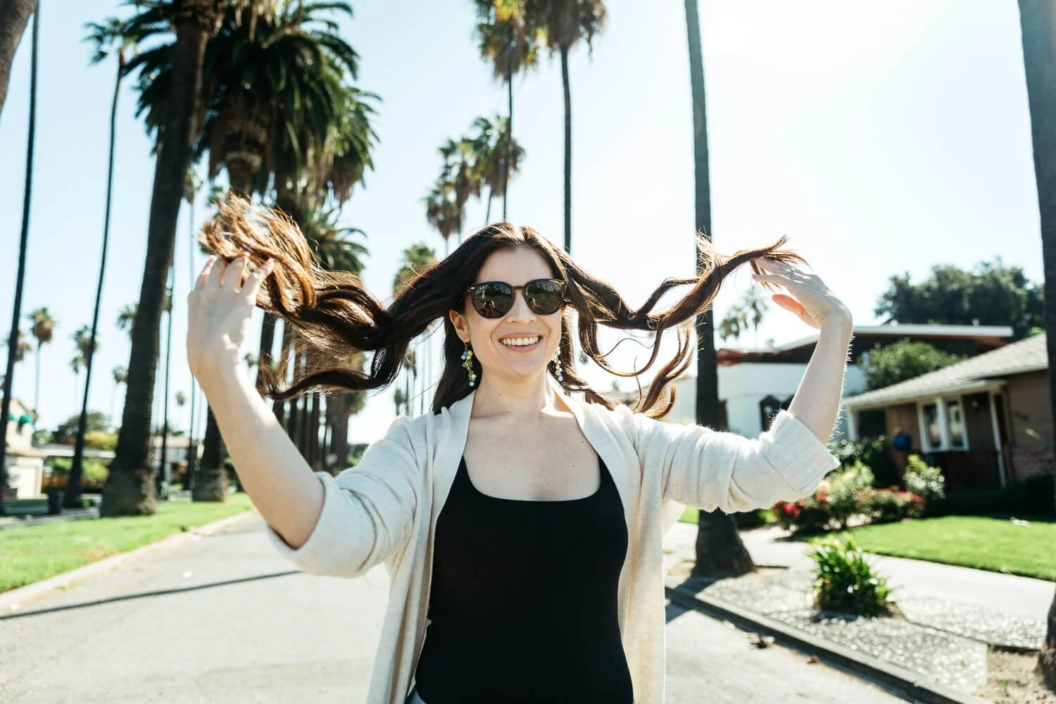 Woman walks down street lined with palm trees while flipping her long brown hair with her hands. Her hair catches the sunlight and she smiles at the camera. The pose keeps her hands busy while looking natural and candid.