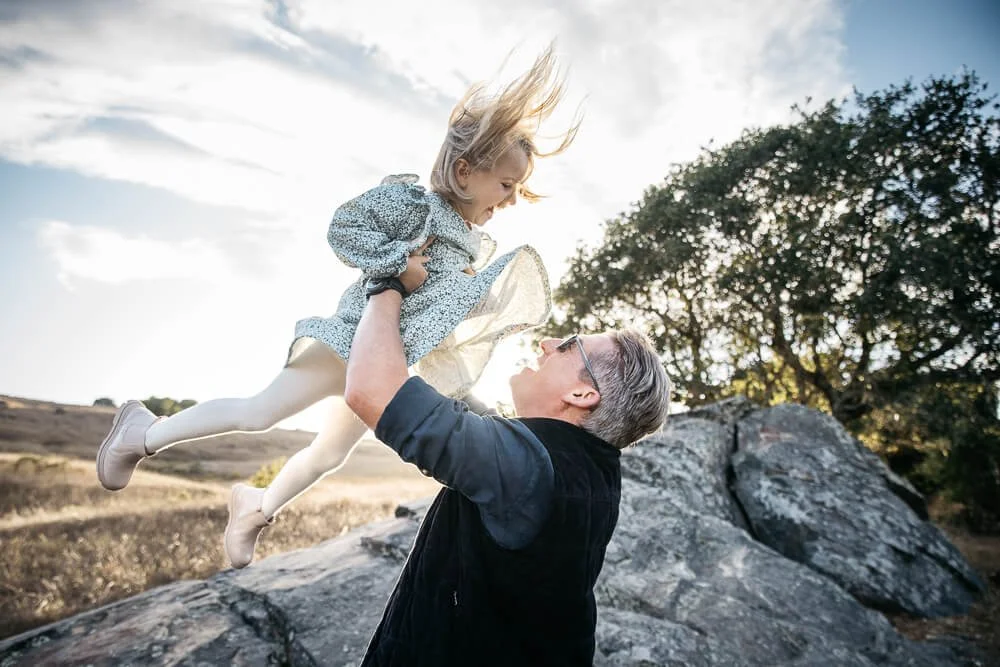Dad wearing sunglasses laughs and tosses young daughter in the air as she smiles down at him, golden hour sunlight streaming in through her dress. Shot at Ring Mountain in Tiburon during outdoor lifestyle family photo shoot.