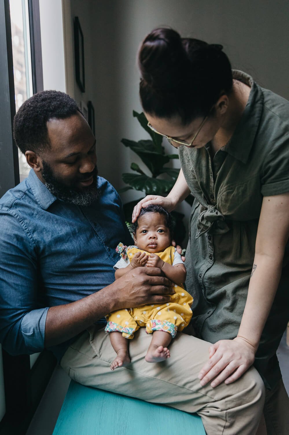 Mom and dad caress newborn baby who wears yellow romper and looks directly at camera during newborn session at home in Oakland.
