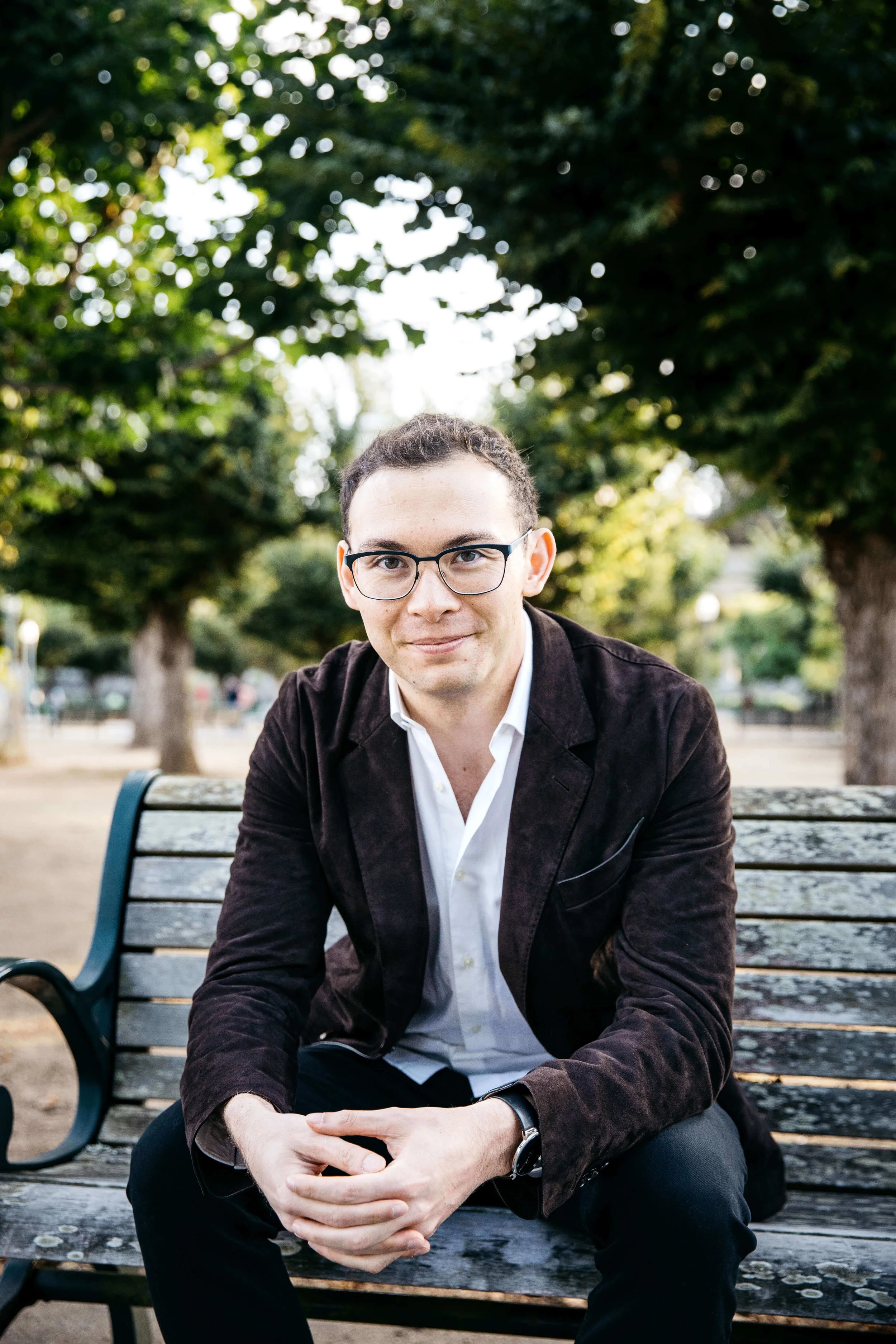 Man wearing glasses and brown jacket, sits on park bench with hands clasped. Shot in Golden Gate Park.