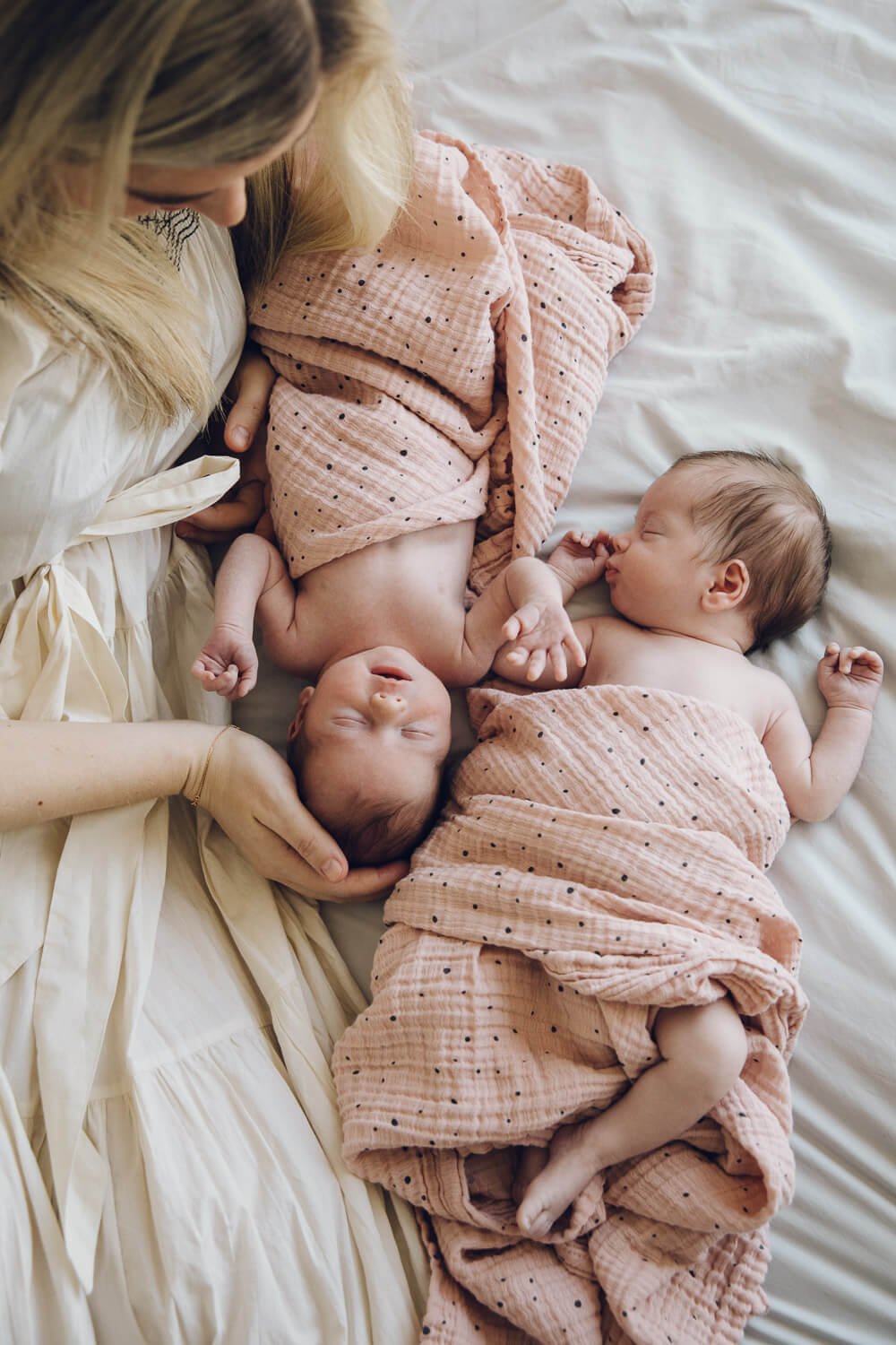 Mom lies on bed and gazes at newborn twins wrapped in pink swaddles, taken at home in Kentfield.
