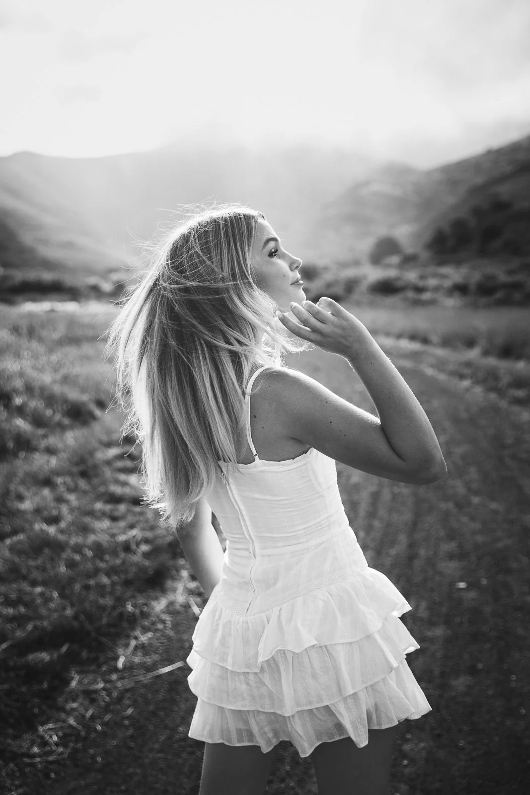 Black and white, backlit shot of teen girl with blond hair and white, ruffled dress standing on a trail with hills in background, shot during senior photo shoot in the Marin Headlands.