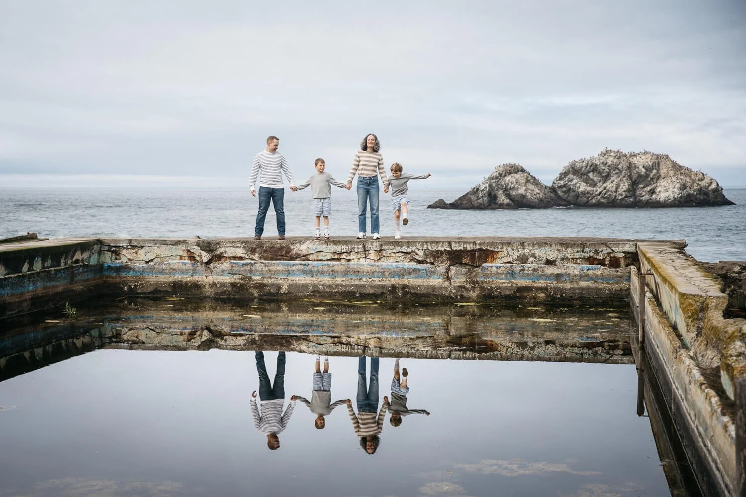 Family of four stand on stone wall at Sutro Ruins, while their reflection shines in water in  front of them.