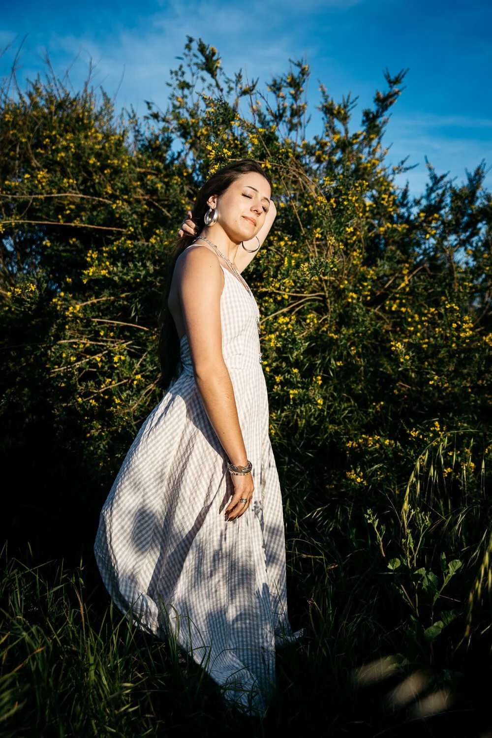San Francisco senior photographer photographs high school senior in a flowing white gingham dress at the Albany Bulb, standing among yellow wildflowers with eyes closed in golden hour light.