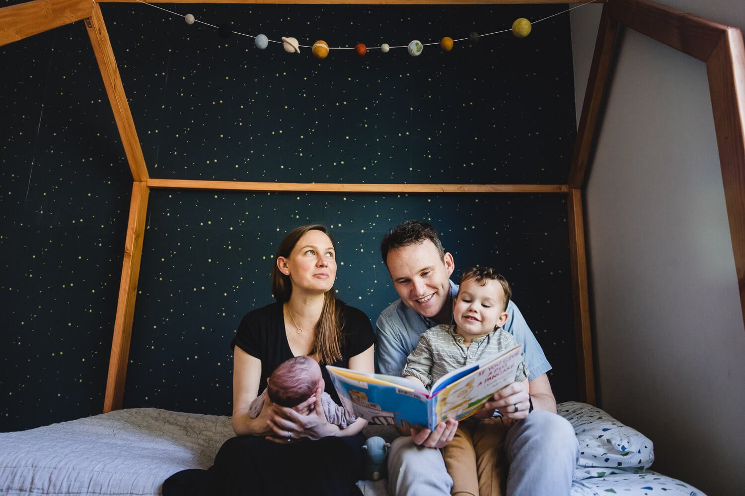 Family sits together on bed while dad reads book to toddler son and mom holds baby during a newborn session at home in San Francisco.