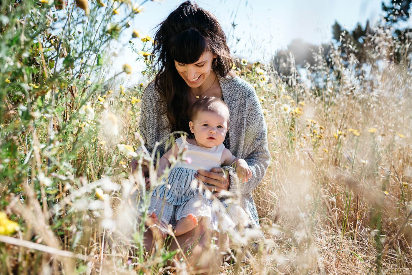 Mom sits in filed of yellow flowers with baby daughter on her lap.