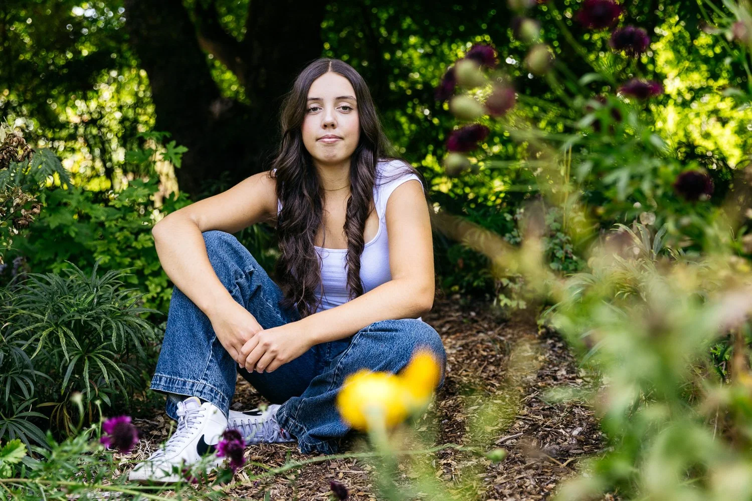 Teen in white tank and jeans sits in garden with purple and orange flowers. Shot during senior photo shoot at Blake Gardens in Berkeley.