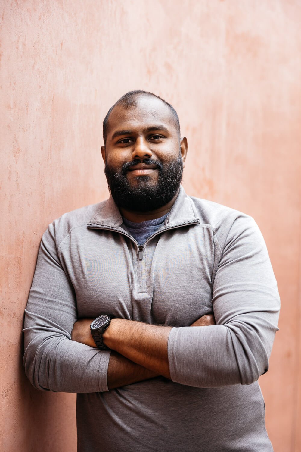 Man in gray half-zip shirt stands with crossed arms against a peach colored wall at the deYoung Museum in San Francisco.