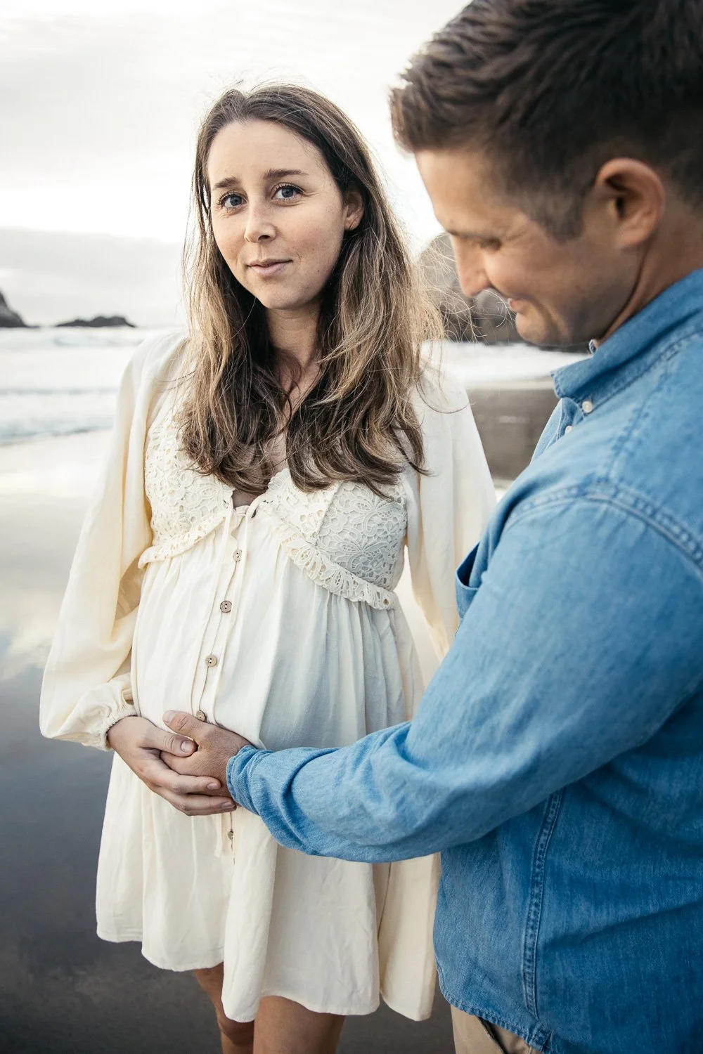 Pregnant mama in white, lacy dress gazes at camera while husband places his hands on her baby bump. They stand in wet sand on the beach as waves roll in behind them.