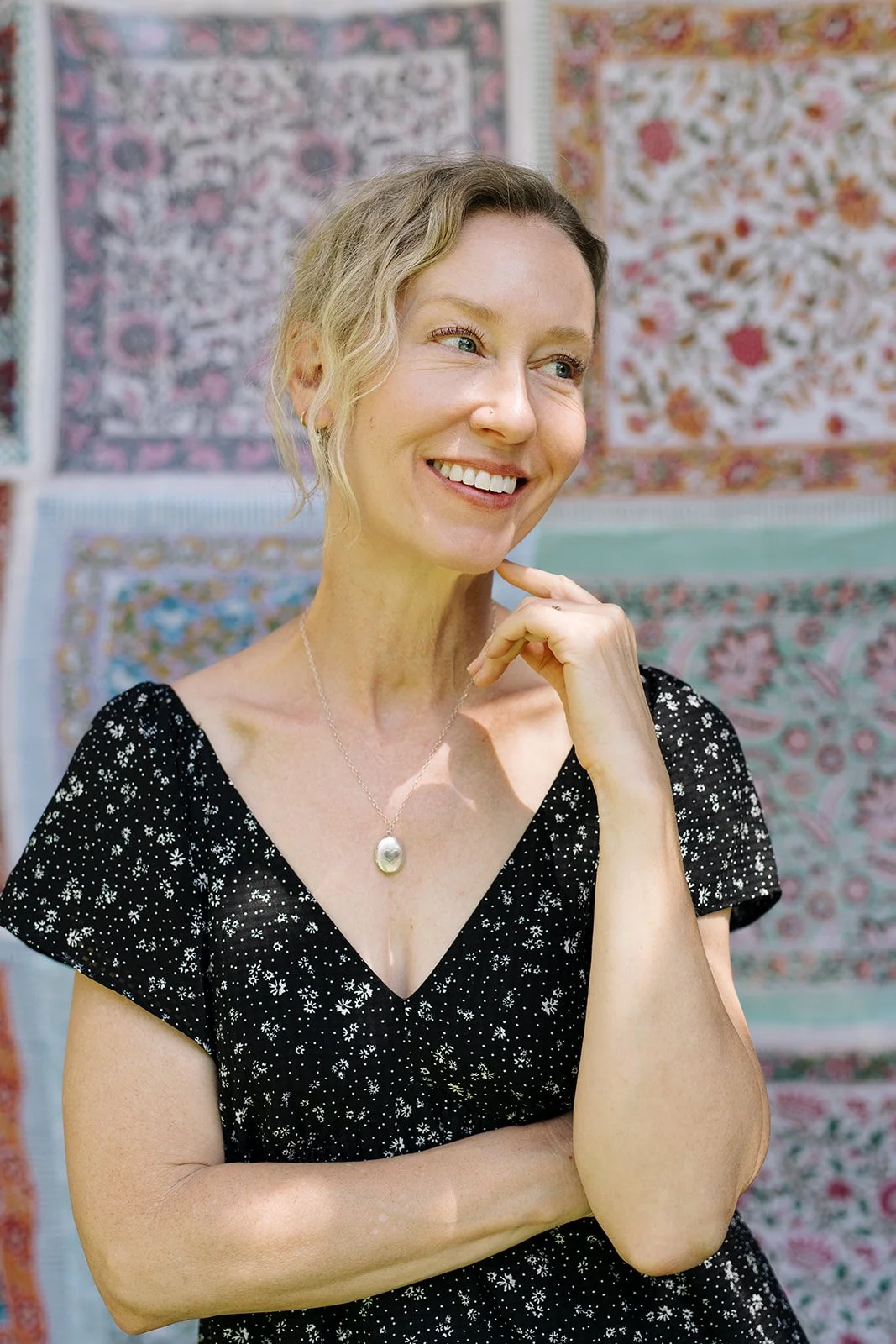 Blond woman wearing black dress with little white flowers, stand in front of flowered wall hanging, with crossed arms and smiles, looking off to the side.