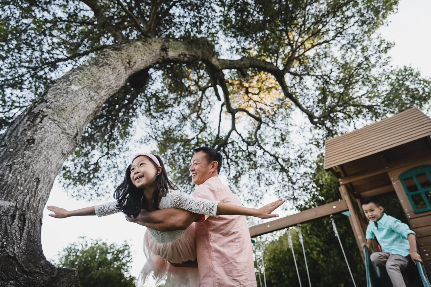 Dad holds young daughter and swings her like an airplane, while standing under a large oak tree. His son plays on the jungle gym behind them. Shot during a family lifestyle photo session in their home backyard.