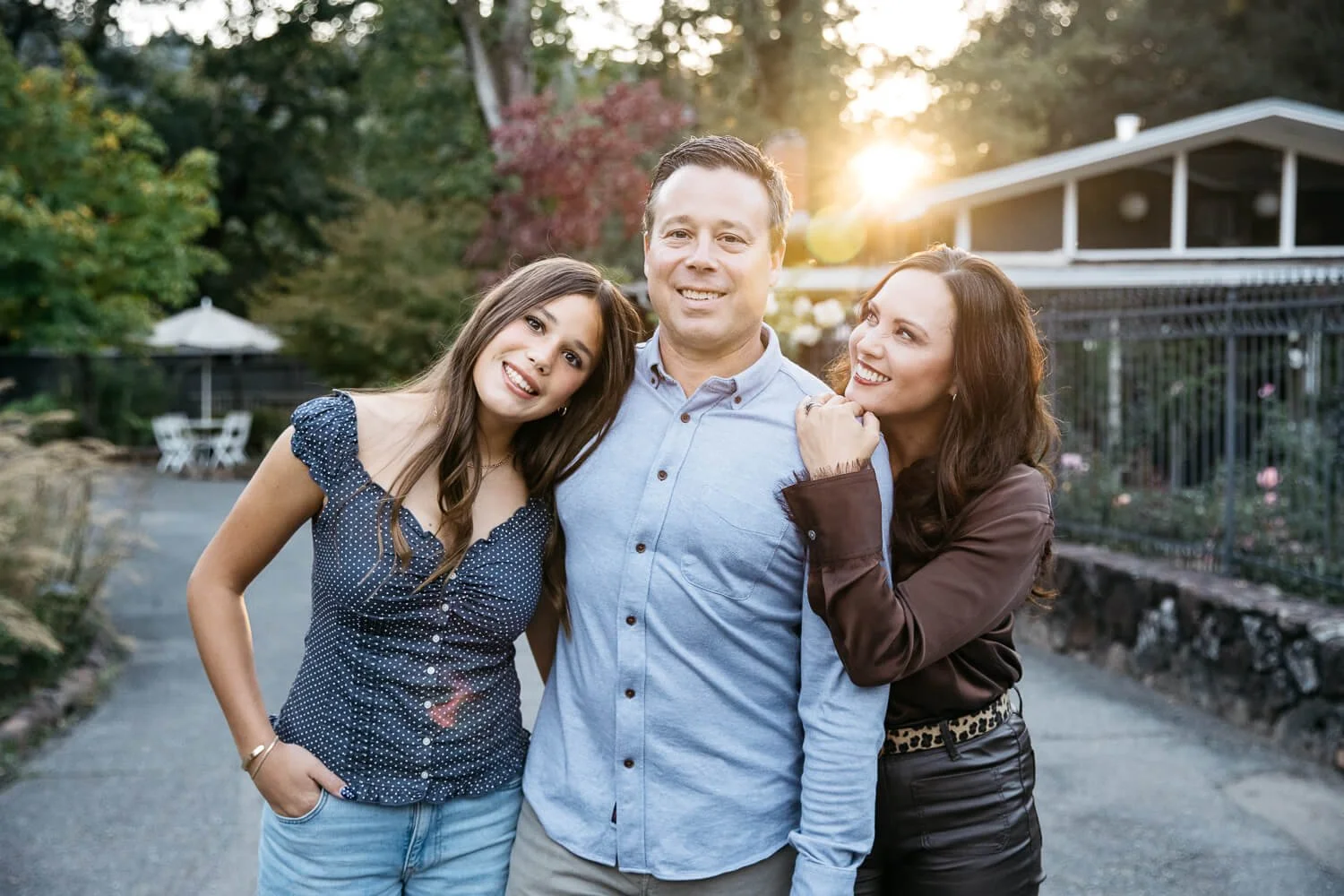 Family of three with teen daugther stand in garden with sunset flare behind them.