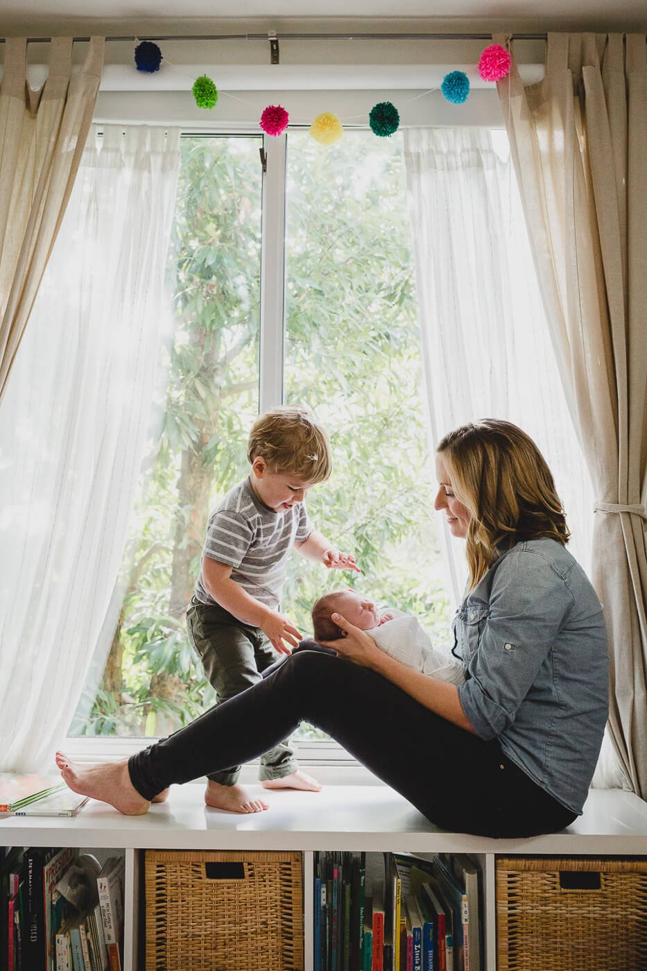 Profile shot of mom sitting at window seat holding baby while toddler brother reaches down to touch baby, taken at home in Pacifica.