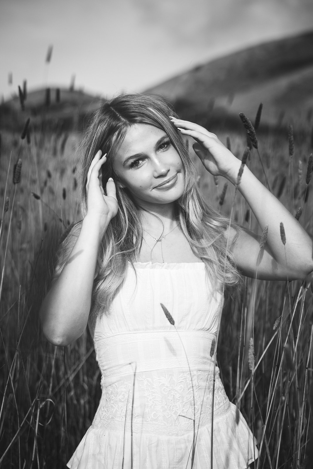 Black and white, front lit shot of teen girl with blond hair and white, ruffled dress standing in tall grasses, shot during senior photo shoot in the Marin Headlands.