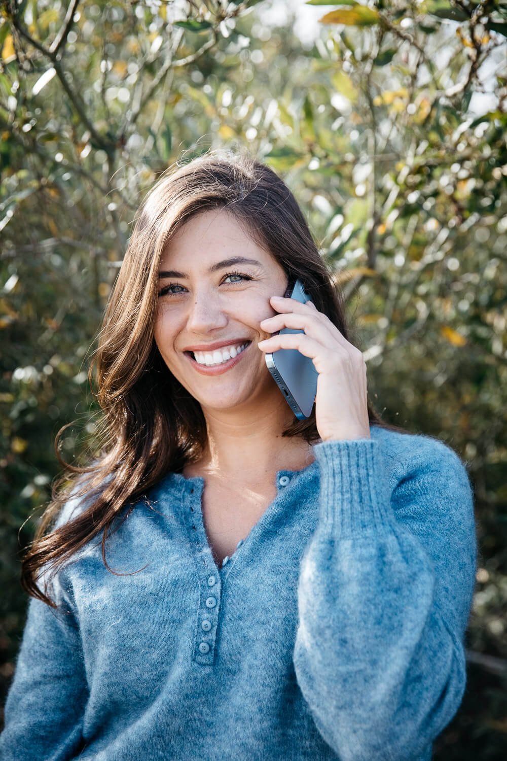 Woman in blue sweater talks on phone and smiles while standing in front of olive bush in the Presidio, San Francisco.