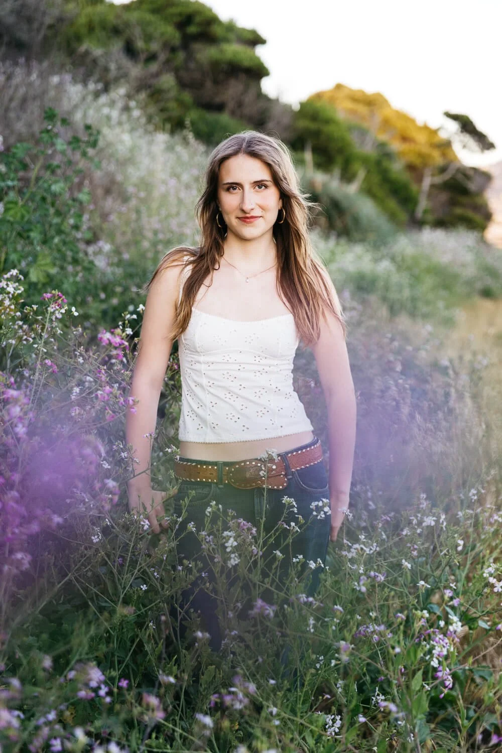 Berkeley senior photographer photographs high school senior standing among purple and white wildflowers at the Albany Bulb, wearing a white eyelet tank top and jeans with a studded western belt in golden hour light.