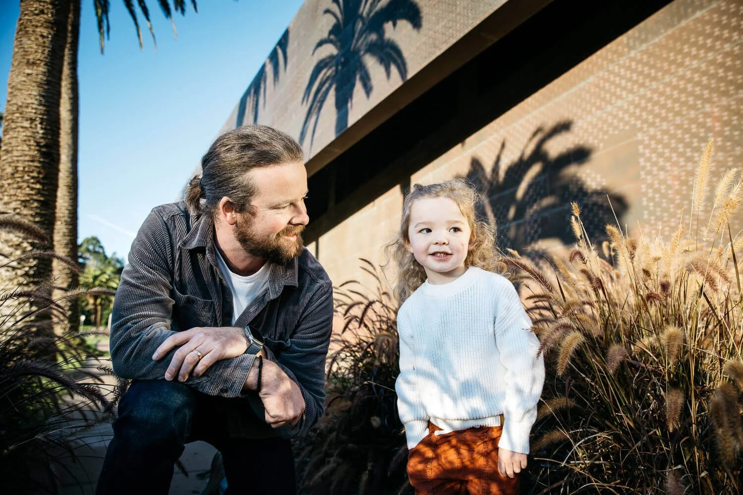 Bearded dad kneels next to toddler son in front of deYoung Museum, with shadows of tall palm trees looming behind them.