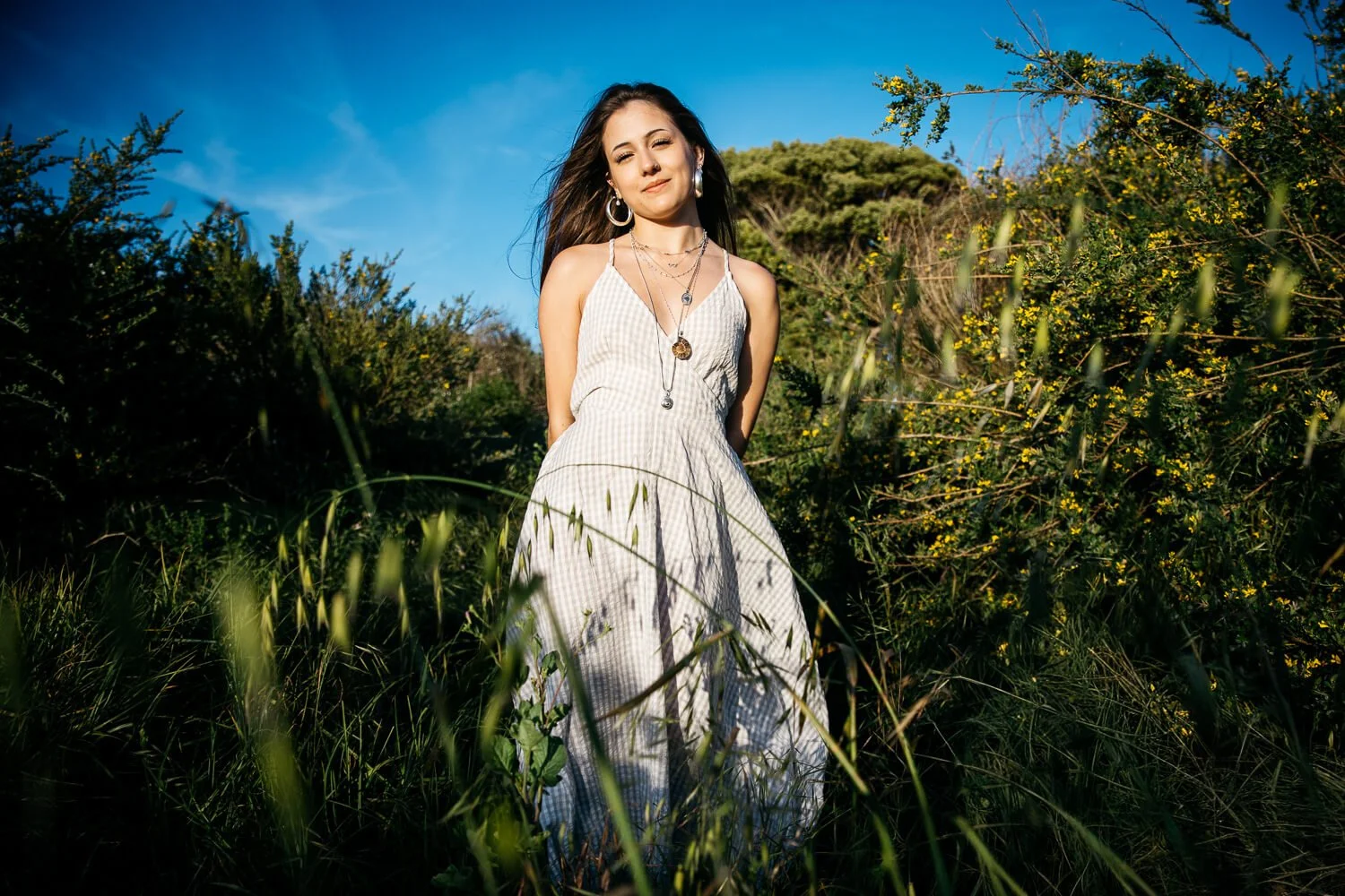 Teen girl in long gingham dress stands in tall grass with wind blowing hair, shot during senior photo session at Albany Bulb.