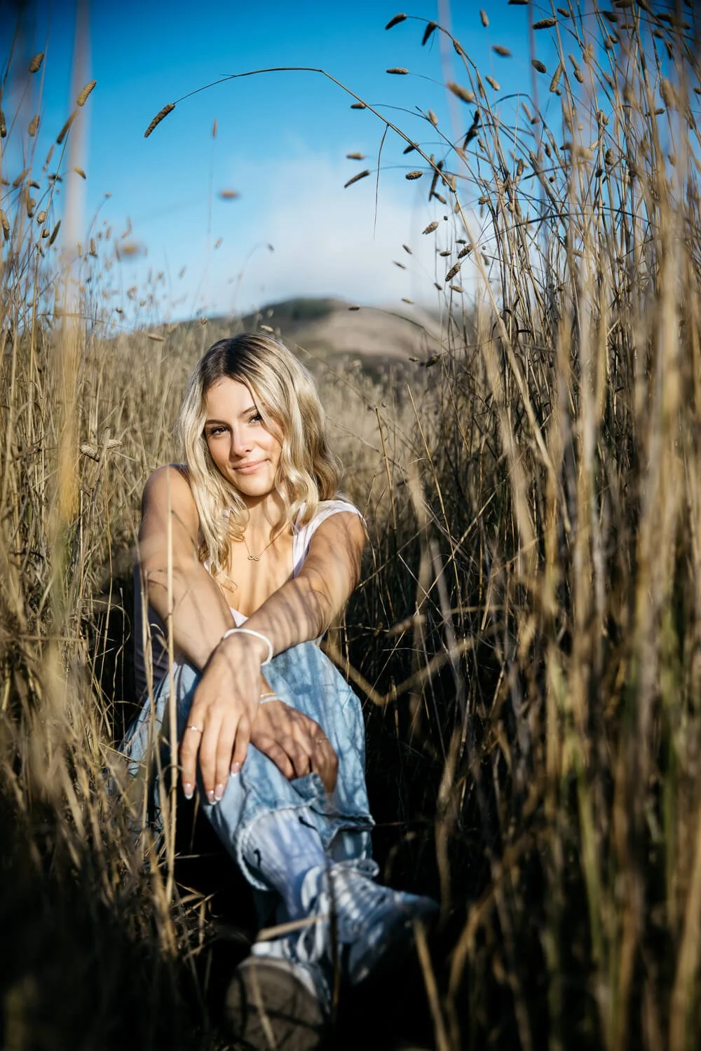 Blond teen kneels in tall golden grass with arms draped over legs, shot during senior photo session in the Marin Headlands.