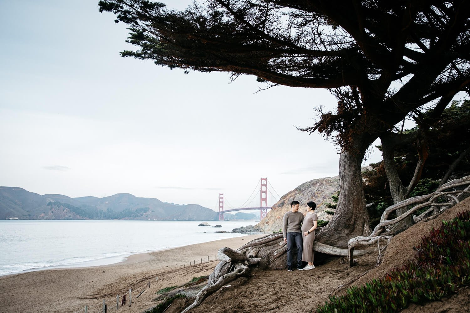 Preganant mama and husband face one another on hillside with Golden Gate Bridge in the background, shot at Baker Beach.