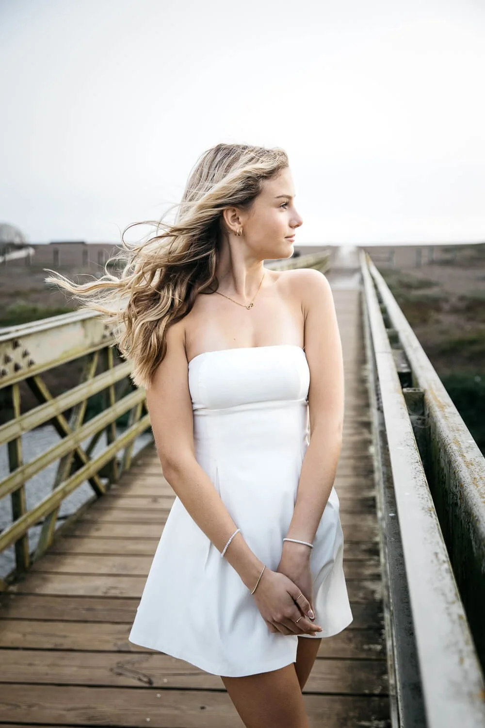 Teen girl with long hair and short white dress stands on footbridge on Rodeo Beach, during senior session.