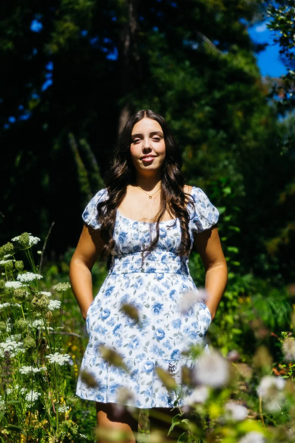 Teen wearing white dress with blue flowers stands in a sunny patch of wildflowers during portrait session in Berkeley.